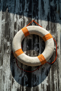 A white and orange circular life preserver hangs on a weathered wooden wall. It is designed as an emergency flotation device for safety in the ocean or pool.