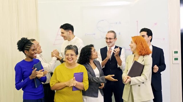Diverse office team applauds during a presentation, standing before whiteboard charts, smiling and celebrating success.