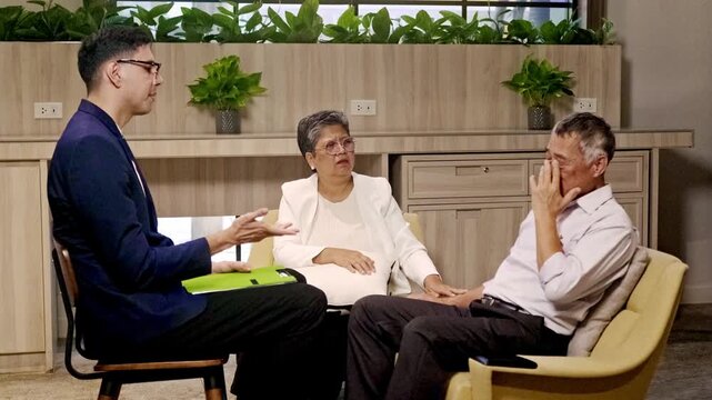 Therapist leads a counseling session with a couple in a modern room; the man wipes tears as the woman offers support amid wooden decor and potted plants