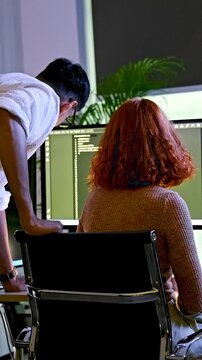 Two coworkers review code on a desktop monitor in a modern office, collaborating on software development as one advises the other.