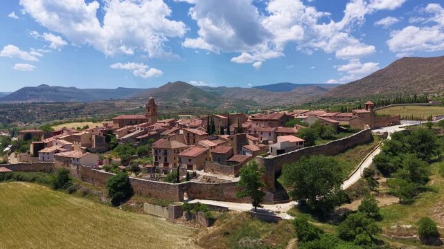 Aerial view of Cantavieja's intact medieval walls and round defensive tower, cypress-lined path and terracotta-roofed houses surrounded by Maestrazgo mountains, Teruel, Aragon, Spain