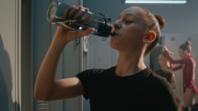 Young Athlete in Black Gymnastics Outfit Stands by Locker, Taking Refreshing Break to Drink Water From Sports Bottle. Girl Gently Stretches Neck, Shoulders, Recovering Energy Before Training Session.