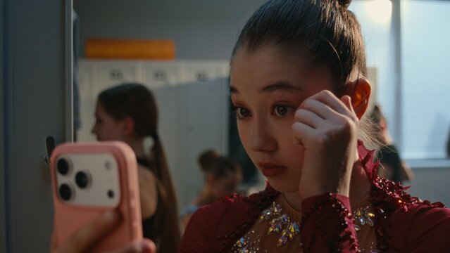 Young Female Uses Smartphone Front Camera as Mirror, Checking Makeup Before Training or Performance. Girl Smiles and Makes Peace Sign For Selfie in Locker Room. Youth Lifestyle, Social Media Culture.