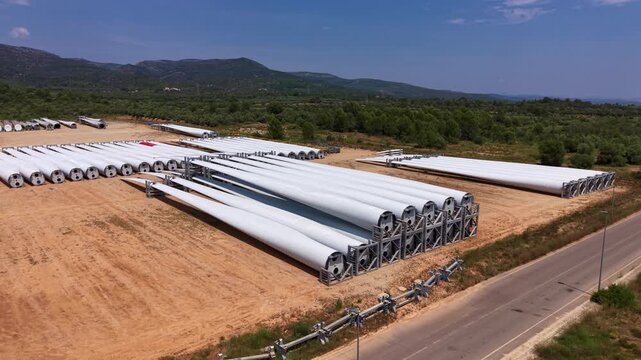 Low oblique aerial view of wind turbine blades arranged in rows at an outdoor storage yard in Maestrazgo region, Teruel, Spain, with forested hills in background