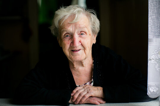 Smiling elderly woman resting her hands on a kitchen table beside a window illuminated by soft natural light in a warm home portrait