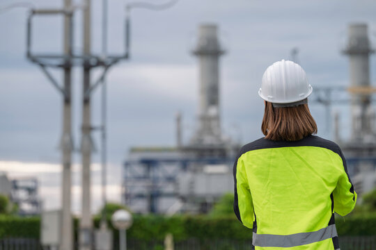 Professional female industrial engineer wearing safety gear uses a clipboard to inspect electrical infrastructure at a modern power plant facility to ensure reliable daily energy production.