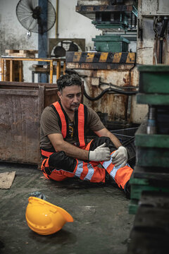 Injured male industrial worker sitting in pain on the floor holding his knee after a workplace accident with a hard hat on the ground inside a manufacturing factory.