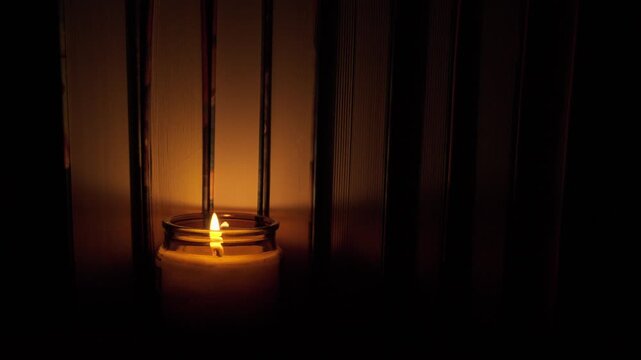 Several books standing in a row with a burning candle nearby on a wooden table. Cozy and warm atmosphere, literature, education, reading, and relaxation in candlelight