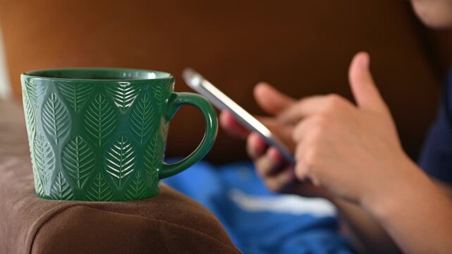 Green mug resting on couch armrest while person scrolls smartphone