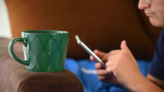 Green ceramic mug resting on couch armrest beside person using phone