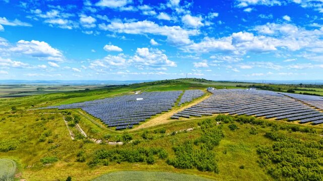 Alternative solar energy farm on green hillside with clouds. Wide angle aerial shot of solar panel rows on grassy green hill under blue sky.