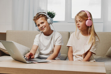 Obraz premium Two children with headphones use laptop at home sitting on couch. Concept of remote learning, entertainment, and digital technology for kids in casual room setting.