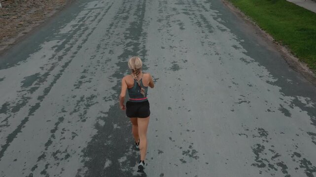 Aerial back view of woman running on open road beside green grass