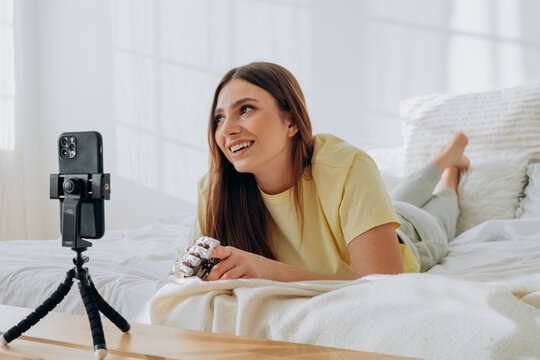 Young woman with a prosthetic bionic hand is lying comfortably on her bed, filming a video for social media platforms using her smartphone on a tripod, sharing her story and lifestyle online