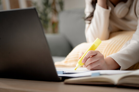 Exam preparation concept. Student highlighting important information while studying with printed documents and a laptop at home for online learning and test revision. Study, exam, highlight, learning,