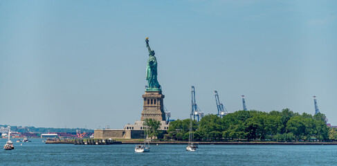 Statue of Liberty and sailboats across New York Harbor. Two yachts pass Liberty Island as container cranes rise behind the green shoreline. © Vadim
