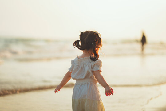 Cute little Asian toddler girl in white dress walking barefoot on the sandy beach shore into the sea wave during a warm golden sunset. Happy child enjoying summer vacation holiday trip, exploring