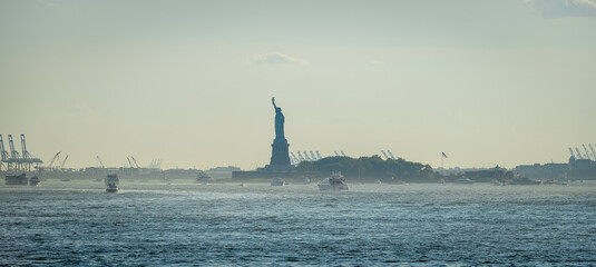 Statue of Liberty standing in hazy New York Harbor. Famous landmark monument on island surrounded by boats in harbor on misty morning, wide panoramic view. © Vadim