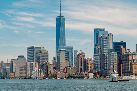 Lower Manhattan skyscrapers with One World Trade Center on sunny day. White coast guard ship and classic sailing vessel cruise in New York Harbor near financial district.