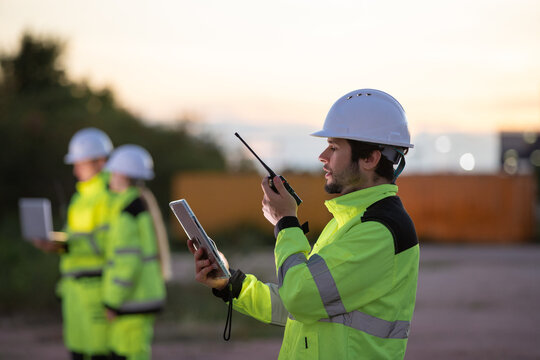 Construction professionals in safety gear collaborating at sunset using tablets and walkie talkies on a busy job site