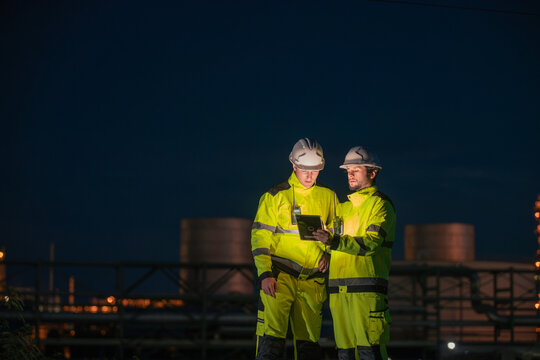 Engineers reviewing data on a tablet at a construction site during twilight with industrial structures in the background