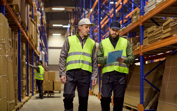 Warehouse workers review inventory logistics on shelves. Two men in safety vests walk aisle racks as a supervisor checks a clipboard to manage orders. Clear roles ensure efficient logistics.
