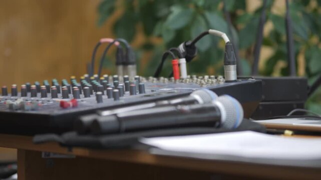 Audio mixing board with microphones on wooden desk. Outdoors, greenery in background. Professional setup for live sound or recording. Ideal for events, workshops, or broadcast scenes