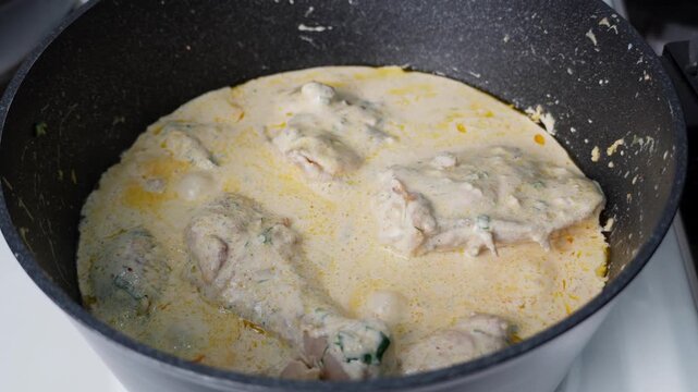Juicy chicken drumsticks simmering in a creamy white sauce with herbs and spices in a deep frying pan. Homemade cooking process, rustic kitchen atmosphere, steam rising from the pan