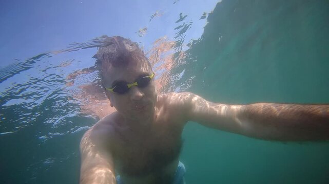 Man wearing goggles making a selfie while swimming underwater in clear blue ocean water during summer vacation