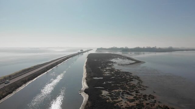 Horizontal 4K 24fps aerial drone video rising and slightly panning to the right over the panoramic Grado lagoon in Italy. The shot shows cars driving on the long causeway bridge road surrounded by blu