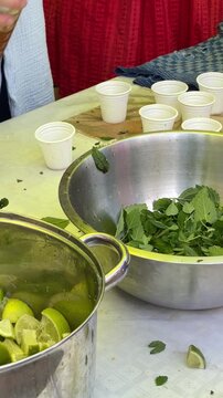 Close-up of hands crushing mint and limes with wooden muddler in translucent cup, preparing beverage samples at tasting station during casual hospitality event