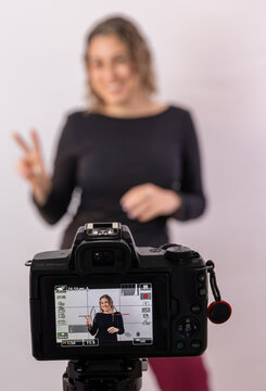 Camera in sharp focus recording a smiling female content creator making a peace sign in the blurred background. Behind the scenes composition representing vlogging, social media, video production and 