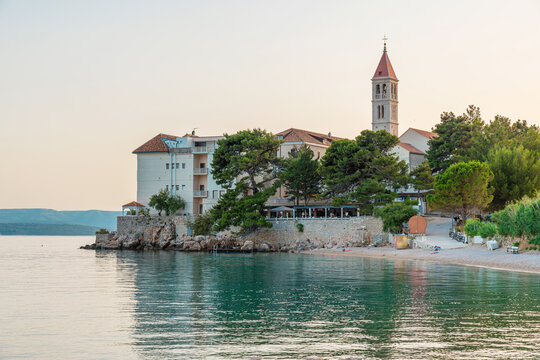 Historic church overlooking calm Adriatic sea water and pebble beach in Bol town on Brac Island, Croatia. Mediterranean coastal architecture, scenic European travel destination, island tourism