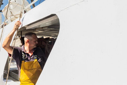 Fisherman in yellow bib overalls on boat deck at harbor