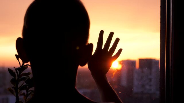 Young child silhouette looking out window at city sunset. Kid putting hand against the glass, creating a sense of longing, hope and contemplative childhood. Back view small boy at sunrise in apartment