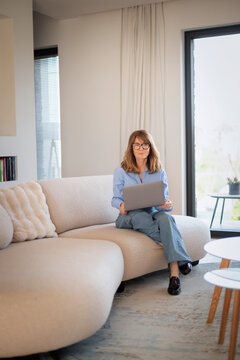 Confident mature woman sitting on a couch in her modern home and using a notebook for work