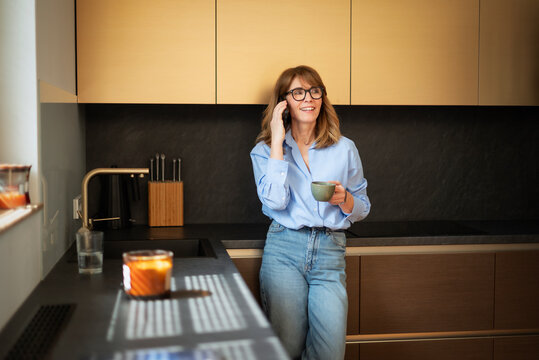 Smiling mature woman with smartphone standing in modern kitchen, enjoying a cup of coffee