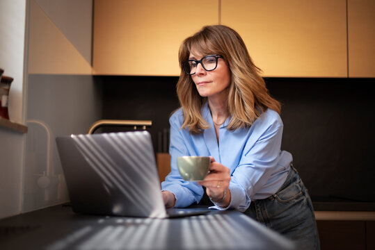 Blond haired smiling woman with laptop standing in a modern kitchen and working from home