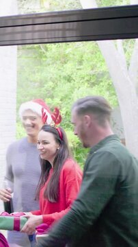 Vertical video: Door opening four adults entering home carrying wrapped gifts in Santa hats hugging