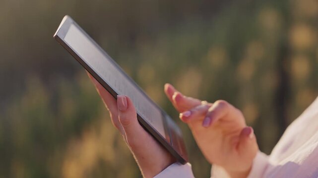 Close-up of hands reading an electronic book on e-reader outdoors, golden hour. Digital reading, modern education, portable library and relaxing lifestyle in peaceful natural setting.