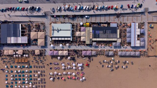 Aerial top down look view of beachfront restaurant cafe complexes with colorful parasols and parked vintage cars on a sandy resort beach under a bright summer sky