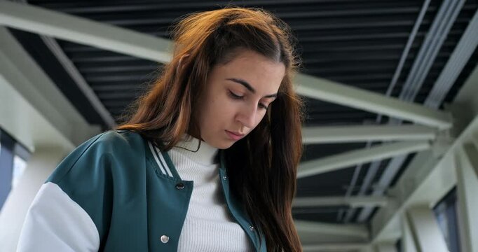 Student in varsity jacket concentrating while typing on colorful mobile device. Dedicated student in casual attire focused on their phone amid modern industrial architectural elements