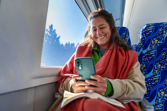 Smiling woman relaxes on train with blanket, book and phone