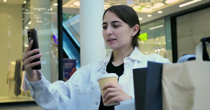 Casual woman sips coffee while shopping. Female customer relaxes with coffee amidst mall shopping scene. Woman in casual attire drinks coffee as she scrolls through her phone at mall