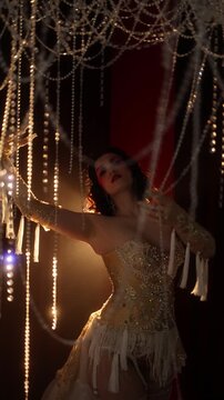 Female showgirl in ornate beaded corset and layered fringe skirt sweeps through dense hanging bead curtain with raised arms, displacing beads and tassels under warm amber backlight, creating