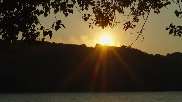 The golden setting sun bursts behind hills above a calm river.