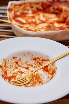 Empty plate with tomato sauce remains and fork in front of baking dish with leftover gnocchi