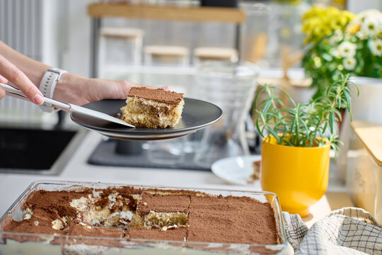 Serving tiramisu slice on dark plate, lifted with spatula from glass dish in kitchen
