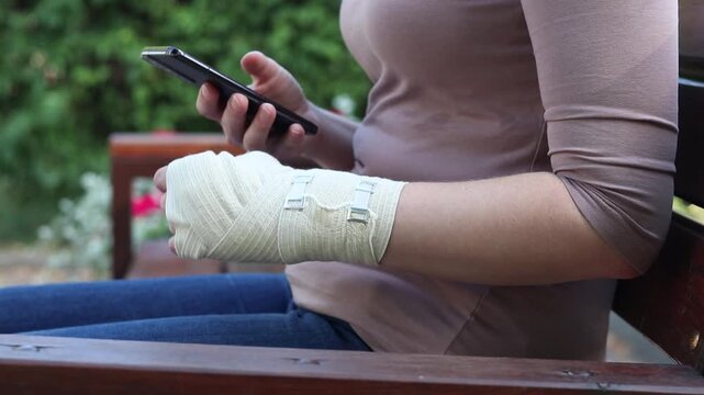 Woman with bandaged hand using smartphone while sitting on park bench. Close-up of woman's hands with elastic bandage on wrist holding mobile phone. Injury and rehabilitation concept