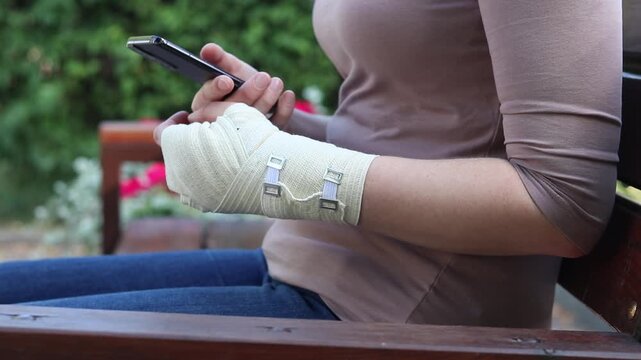 Woman with bandaged hand using smartphone while sitting on park bench. Close-up of woman's hands with elastic bandage on wrist holding mobile phone. Injury and rehabilitation concept